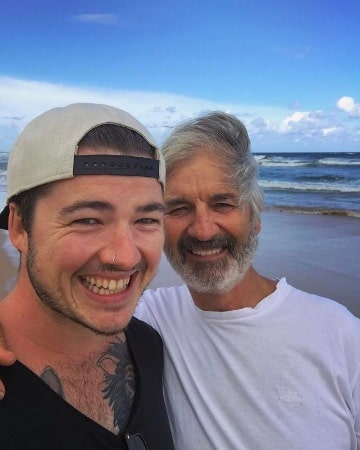 A smiling picture of John Jarratt in white t-shirt with his son Charlie Jarratt with a beautiful ocean background view. 